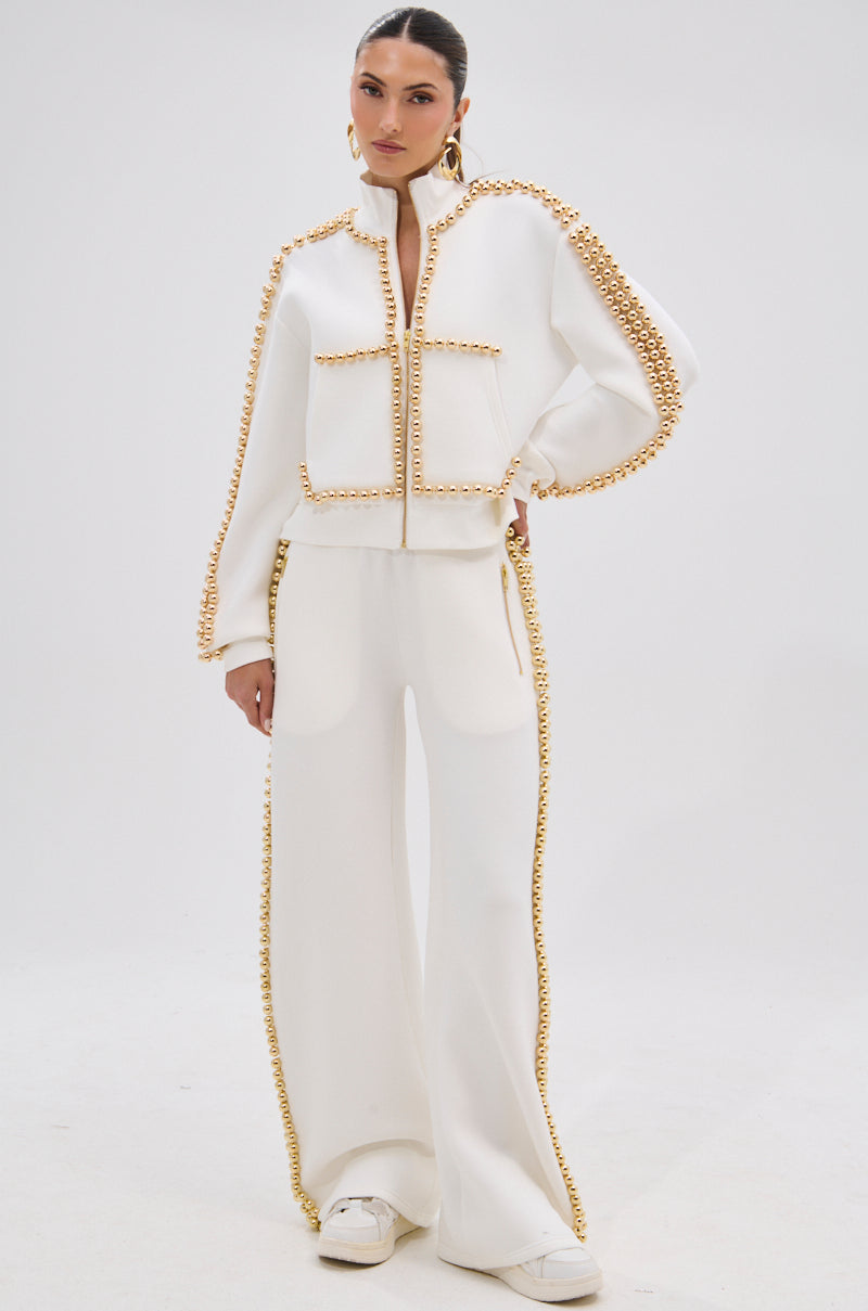 A woman exudes confidence in the BALLS TO THE WALL JOGGER IN WHITE, featuring metallic gold-tone ball embellishments. She styles it with white sneakers, large hoop earrings, and a sleek ponytail for a modern look.