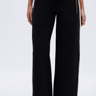 A person wearing the PEARL GIRL JOGGER—black wide-leg sweatpants with zipper pockets and pearl details along the side seams—paired with black open-toe sandals. The photo is cropped from the shoulders down.