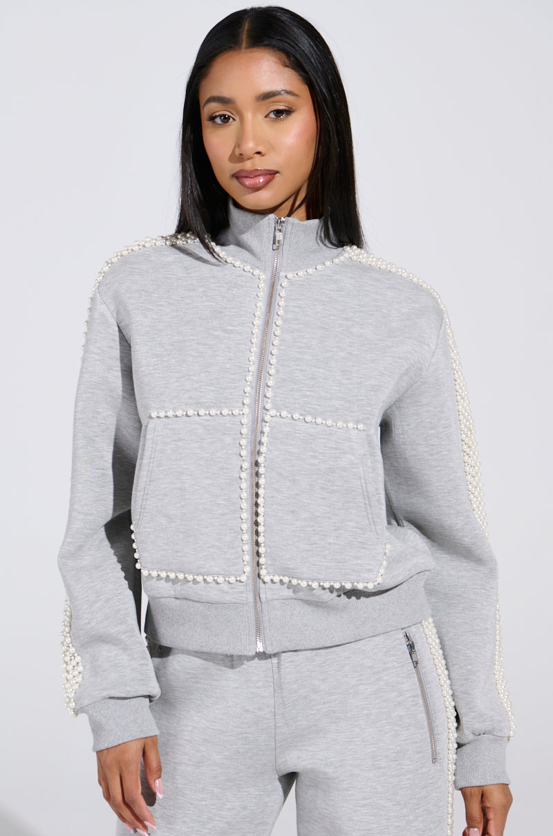 A woman models the PEARL GIRL ZIP UP SWEATSHIRT IN GREY, featuring pearl trim and a zippered front. This high-collared piece is paired with matching pants against a plain backdrop, accented by her straight hair and neutral expression for a chic athleisure look.