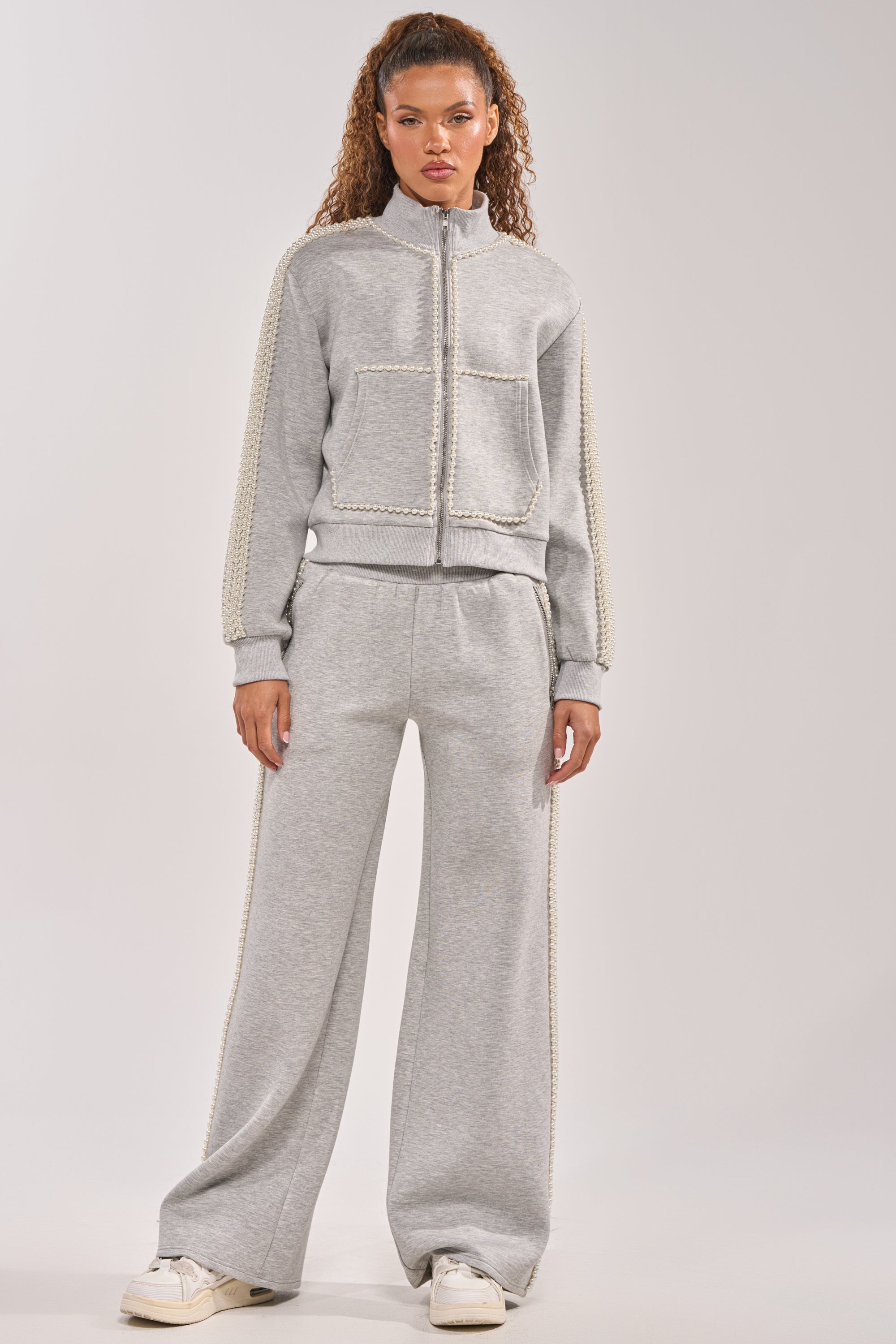 A woman wears the PEARL GIRL ZIP UP SWEATSHIRT IN GREY with front pockets and textured sleeves, paired with matching wide-leg pants and white sneakers. 9