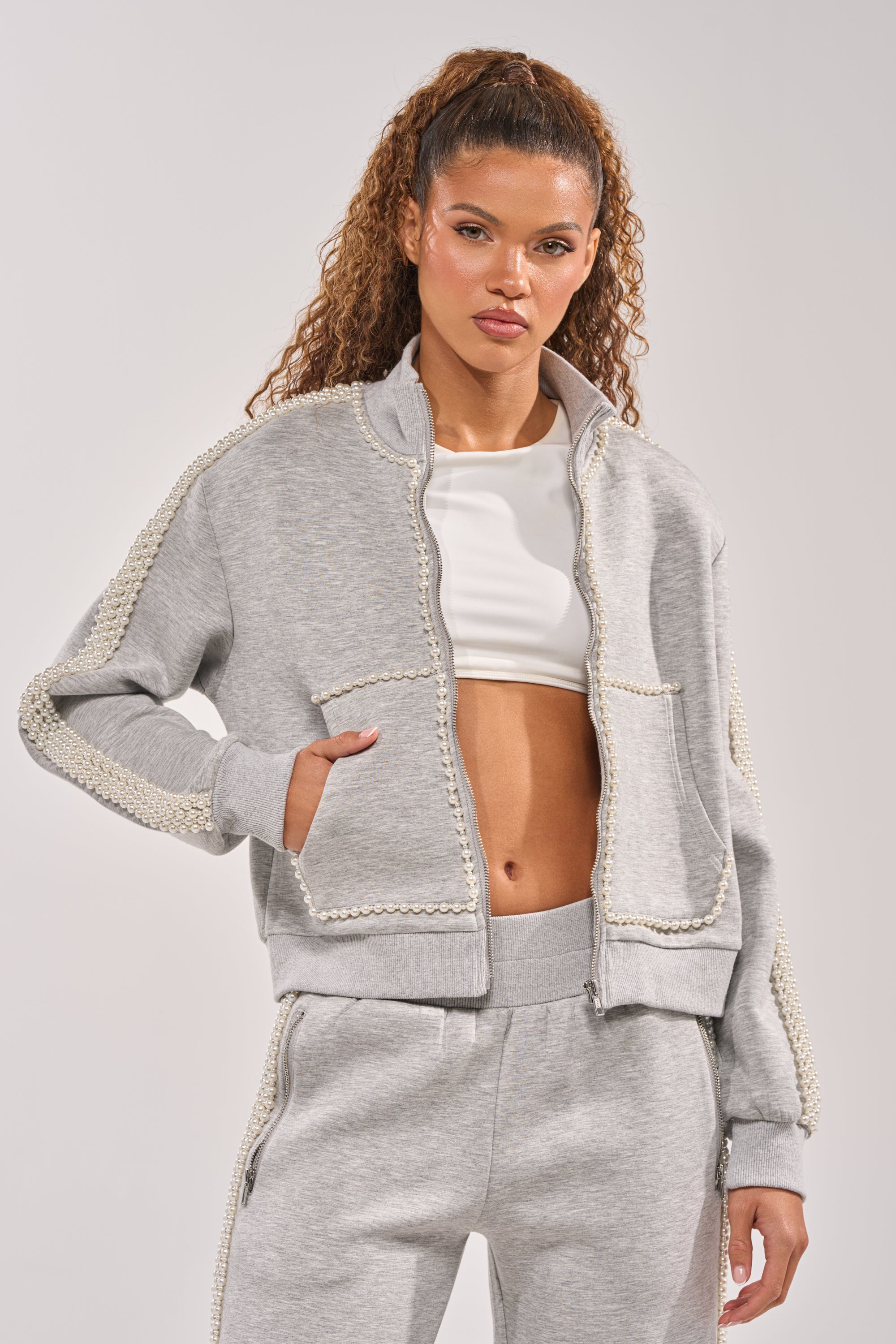 A woman with curly hair models the PEARL GIRL ZIP UP SWEATSHIRT IN GREY, featuring pearl details, over a white crop top and matching sweatpants, posing with one hand in her jacket pocket against a plain background. 0