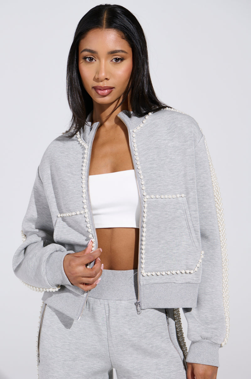 A woman with long black hair wears a PEARL GIRL ZIP UP SWEATSHIRT IN GREY and matching pants. Her jacket is open, revealing a white crop top beneath. With confidence in her expression, she stands against a light background.