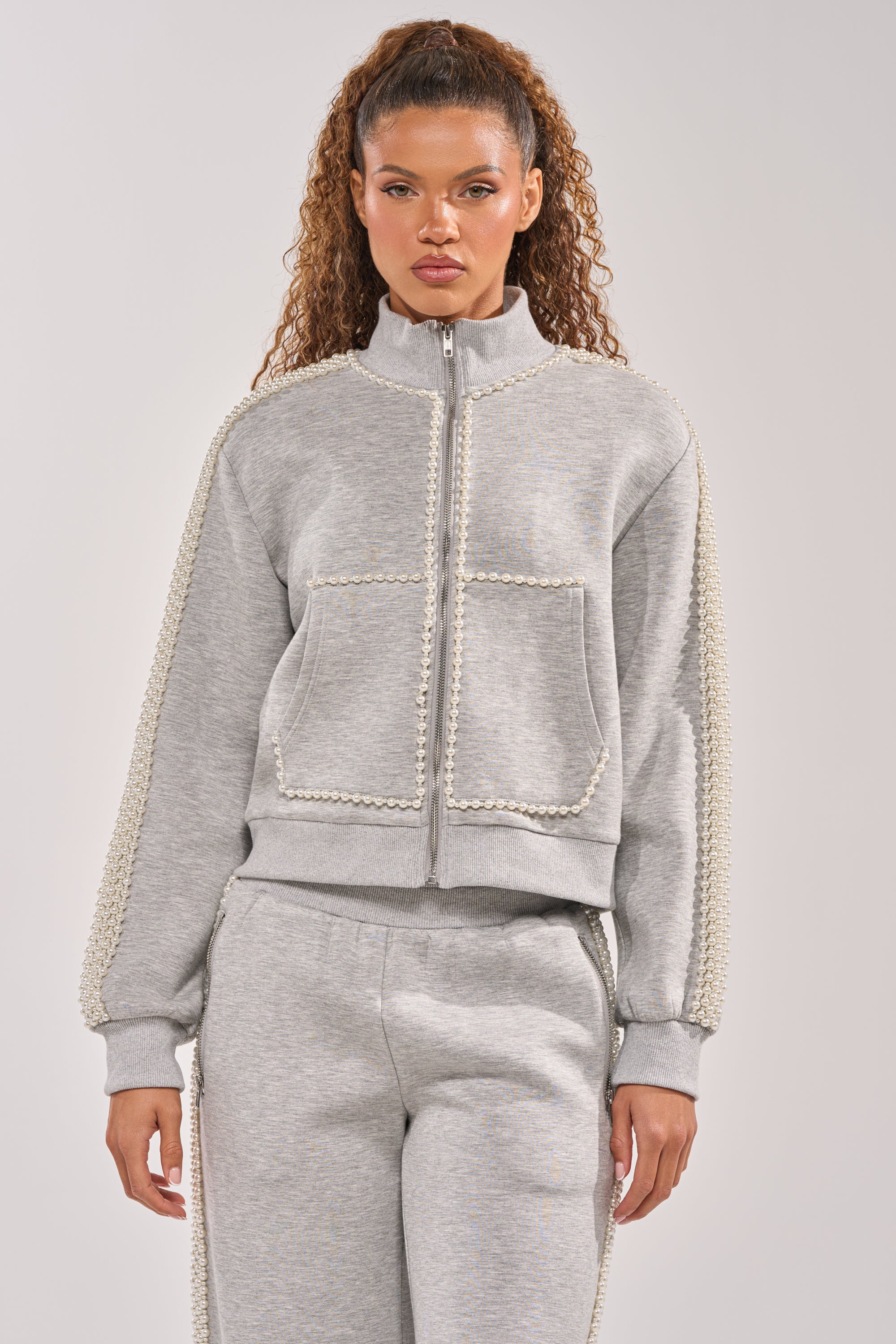 A woman with long, curly hair wears the oversized PEARL GIRL ZIP UP SWEATSHIRT IN GREY with textured trim and matching grey sweatpants, standing against a plain background. 2