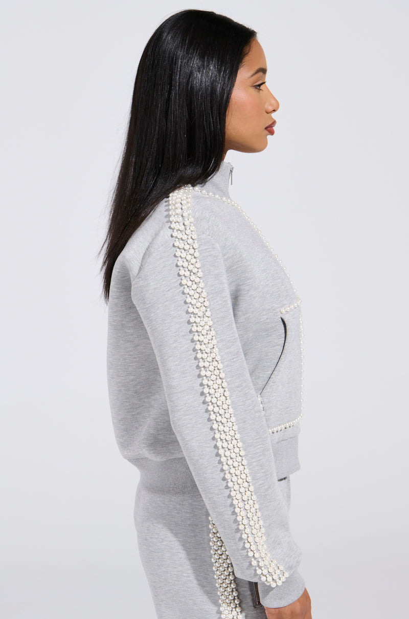 A person with long black hair is wearing the PEARL GIRL ZIP UP SWEATSHIRT IN GREY, featuring pearl-like trims on the sleeves and pockets, standing in profile against a plain background.