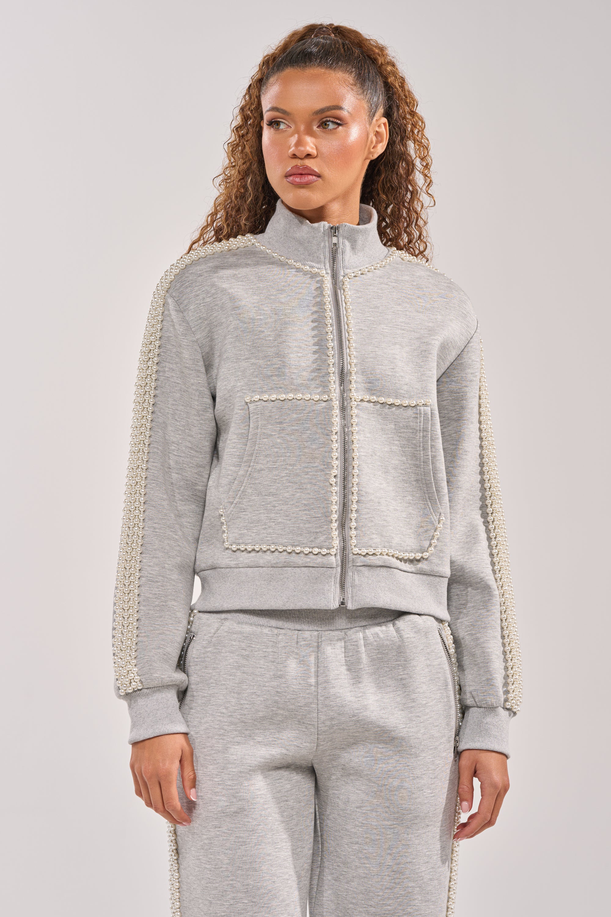 A woman with curly hair wears the PEARL GIRL ZIP UP SWEATSHIRT IN GREY, an oversized tracksuit top with decorative trim on the sleeves and pockets, standing against a plain light background. 3