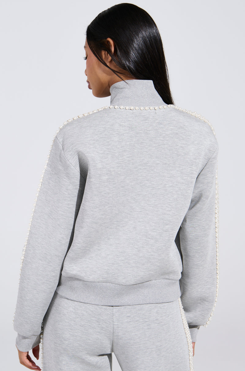 A person with long dark hair faces away, wearing the athleisure-inspired PEARL GIRL ZIP UP SWEATSHIRT IN GREY. The sweatshirt's pearl-like trim along the shoulders and sleeves adds elegance to their sporty look.