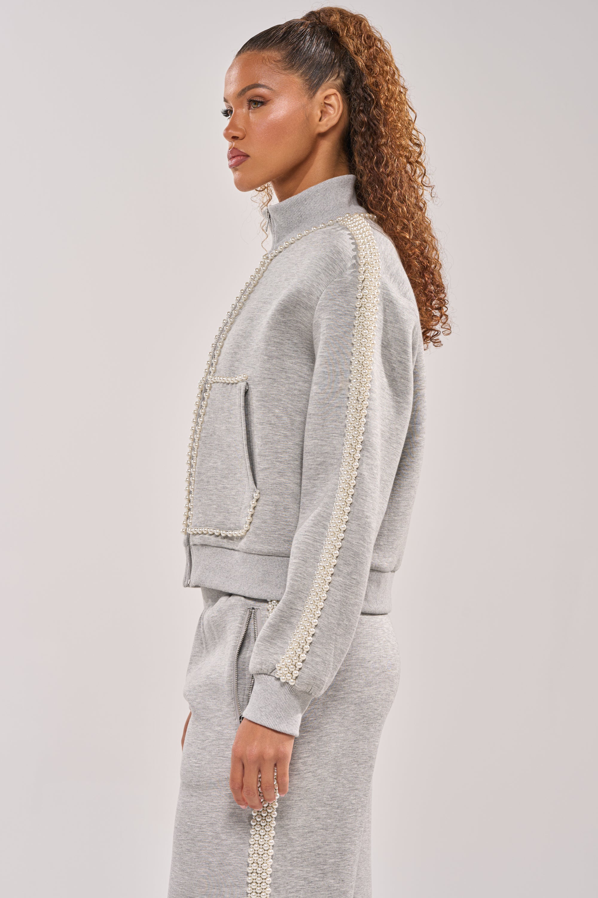 A woman with curly hair in a high ponytail models the PEARL GIRL ZIP UP SWEATSHIRT IN GREY, featuring pearl details on the sleeves and pockets, posed in profile against a plain light background. 4