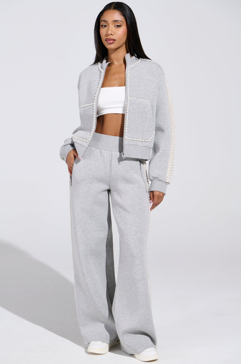 In a PEARL GIRL ZIP UP SWEATSHIRT IN GREY with matching wide-leg pants over a white crop top, she radiates athleisure style against a light background. With hands in her pockets, she completes the chic look with white sneakers featuring elegant pearl trim.