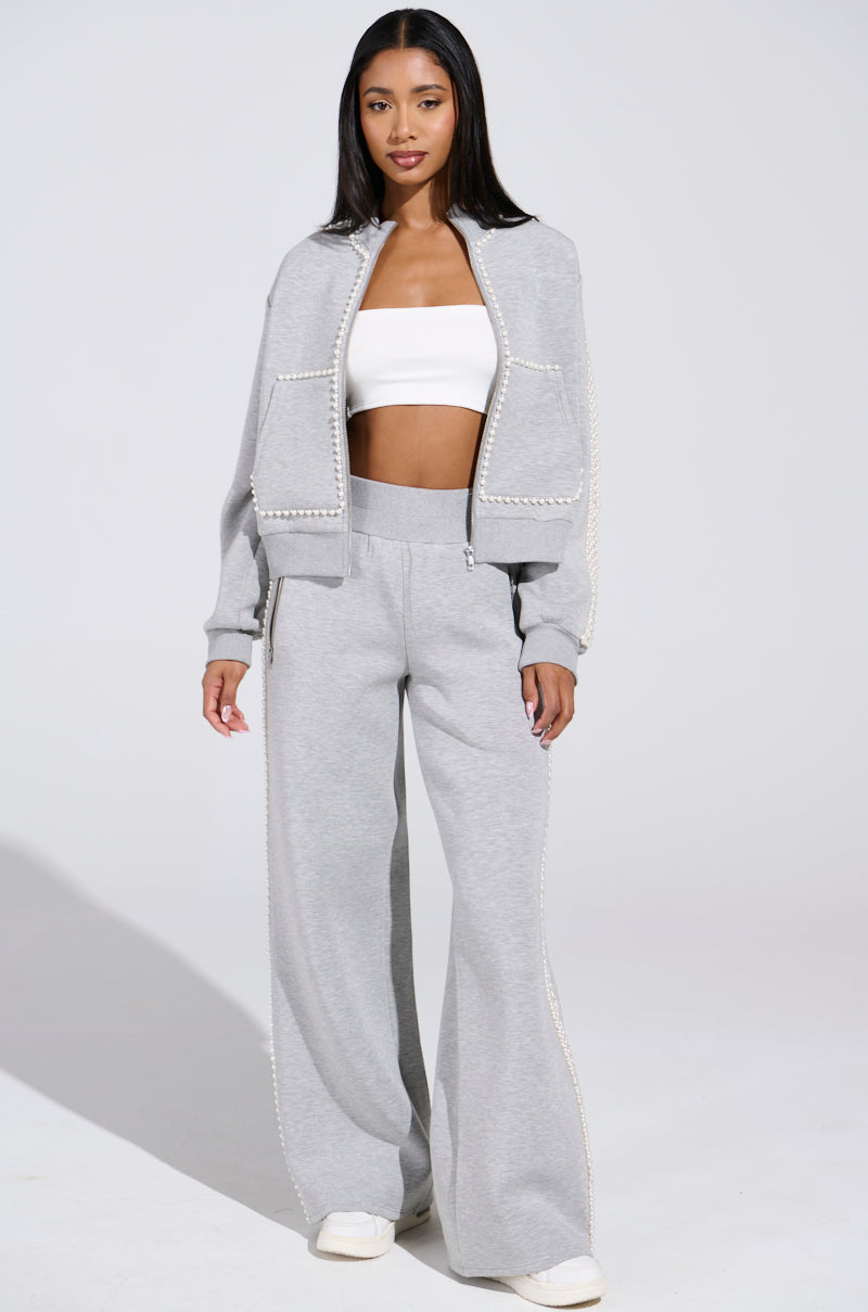 A woman exudes confidence in the PEARL GIRL ZIP UP SWEATSHIRT IN GREY, a light gray tracksuit with wide-leg pants and white trim. Underneath, she wears a white tube top and has long straight hair. Her look is finished with chic pearl-trimmed white shoes.