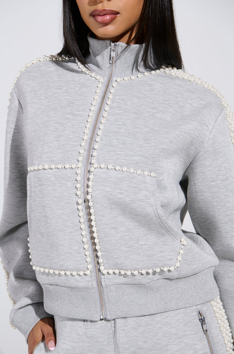 Wearing the PEARL GIRL ZIP UP SWEATSHIRT in grey with pearl-trimmed seams and edges, paired with matching pants, this person exudes an elegant athleisure vibe. The focus is on the upper body and part of the face against a plain, neutral backdrop.