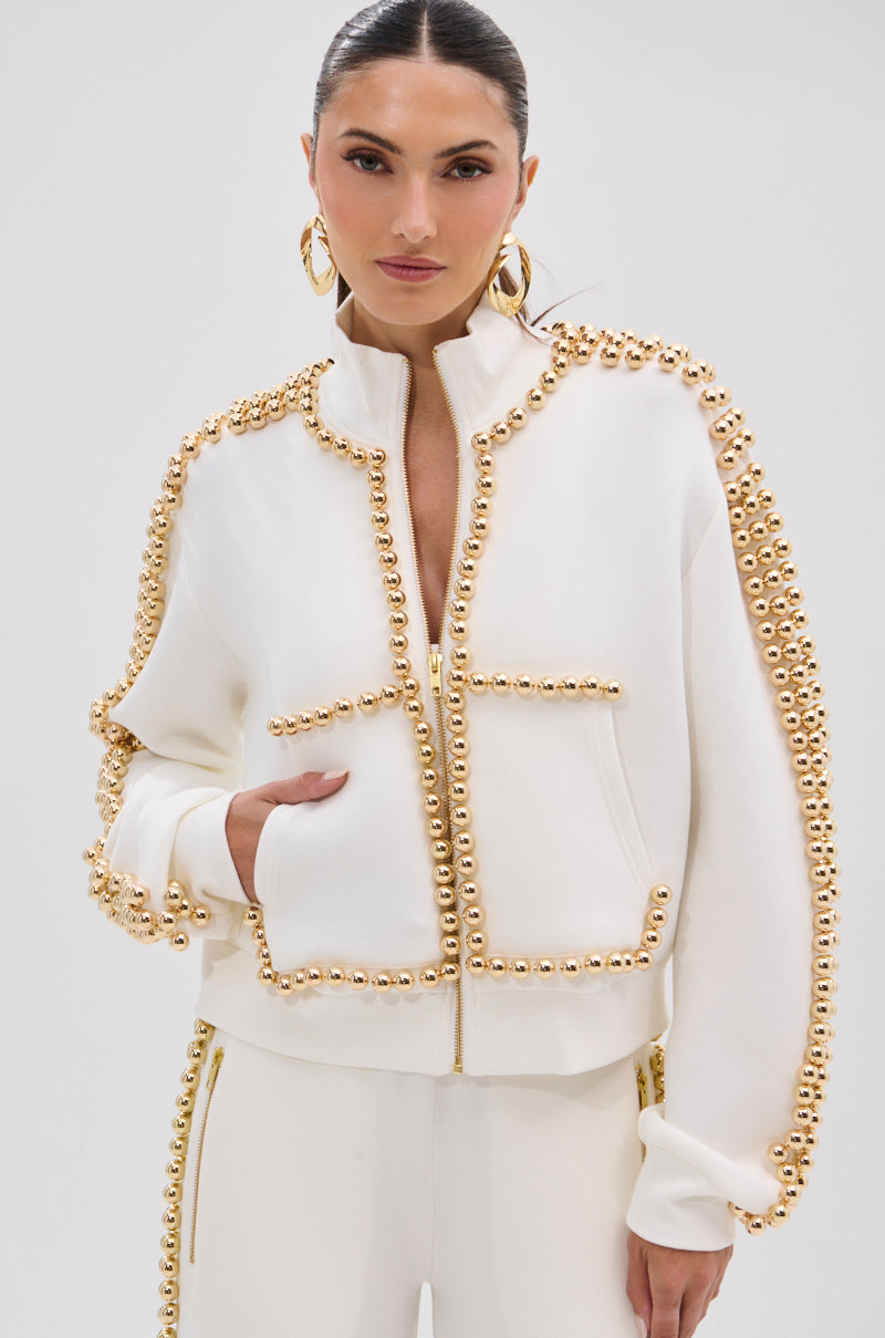 A woman stands confidently against a plain background, wearing the BALLS TO THE WALL ZIP UP SWEATSHIRT IN WHITE featuring metallic gold-tone embellishments, paired with matching pants, gold hoop earrings, and slicked-back hair.