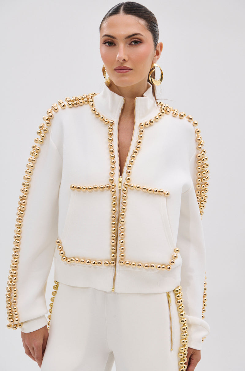 Wearing the BALLS TO THE WALL ZIP UP SWEATSHIRT IN WHITE, a woman shows off metallic gold-tone embellished seams and pockets, paired with matching pants, slicked-back hair, gold hoops, and a bold, confident expression.