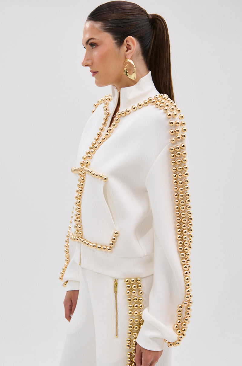 A woman in profile with long brown hair in a ponytail and large gold hoop earrings models the BALLS TO THE WALL ZIP UP SWEATSHIRT IN WHITE, featuring metallic gold-tone embellishments, against a plain light background.