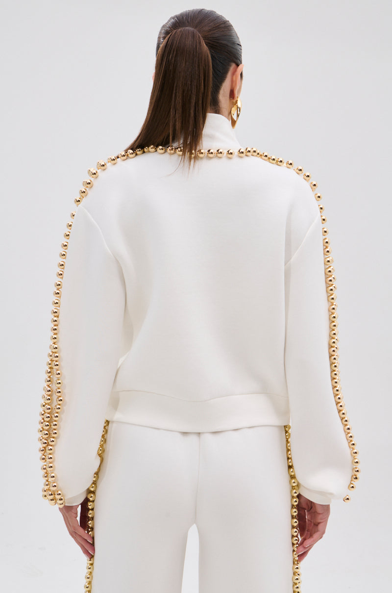 A woman with long brown hair in a ponytail is seen from behind wearing the BALLS TO THE WALL ZIP UP SWEATSHIRT IN WHITE, adorned with metallic gold-tone details on the shoulders and sleeves, against a plain background.