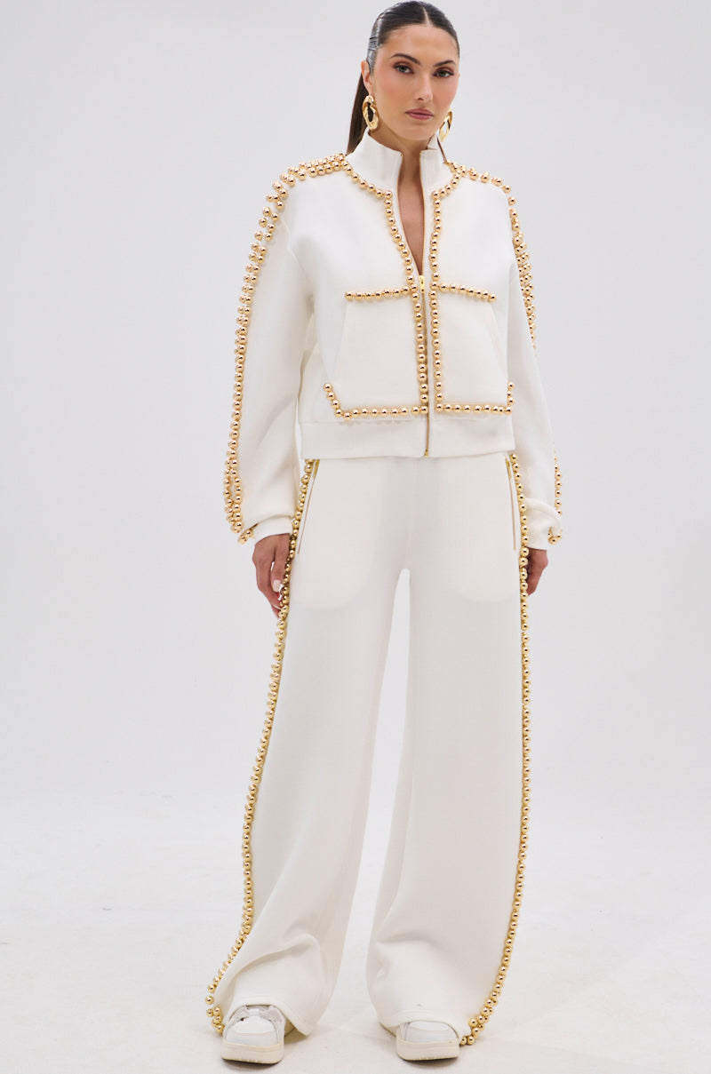A woman faces forward in the BALLS TO THE WALL ZIP UP SWEATSHIRT IN WHITE with metallic gold-tone accents along the seams and sleeves. She wears sleek hair, large hoop earrings, and white shoes against a plain white background.