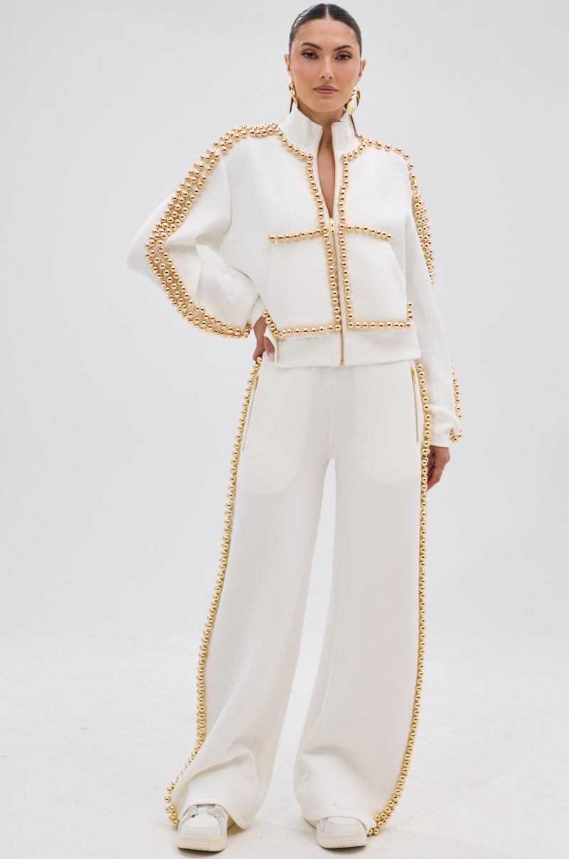 A woman wears the BALLS TO THE WALL ZIP UP SWEATSHIRT IN WHITE, styled forward with wide-leg pants, metallic gold-tone seam details, large front pockets, white shoes, and gold hoop earrings.