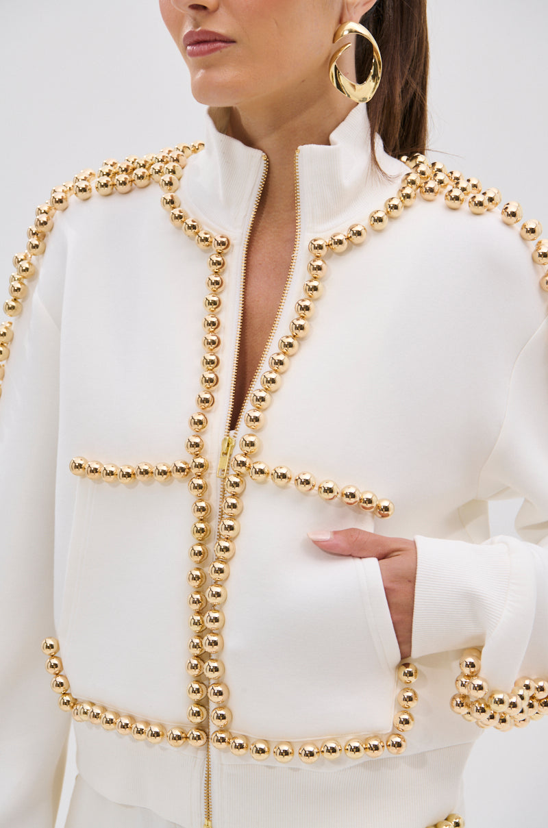 A woman wears the BALLS TO THE WALL ZIP UP SWEATSHIRT IN WHITE, featuring metallic gold-tone embellishments along the seams, with one hand in her pocket. She accessorizes with large round gold hoop earrings; her face is partly visible.