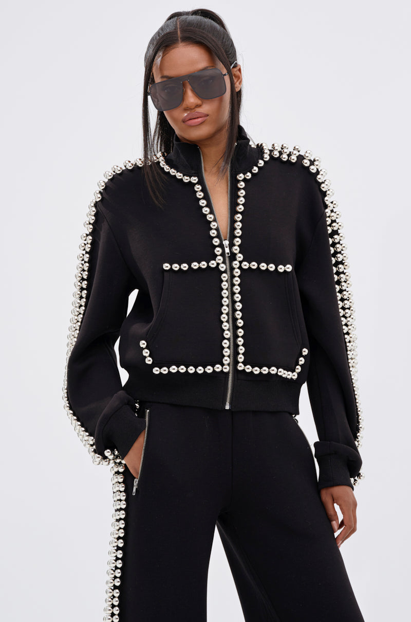 A woman wears the BALLS TO THE WALL ZIP UP SWEATSHIRT IN BLACK, detailed with oversized silver ball embellishments, paired with matching pants, oversized sunglasses, and sleek half-up hair.