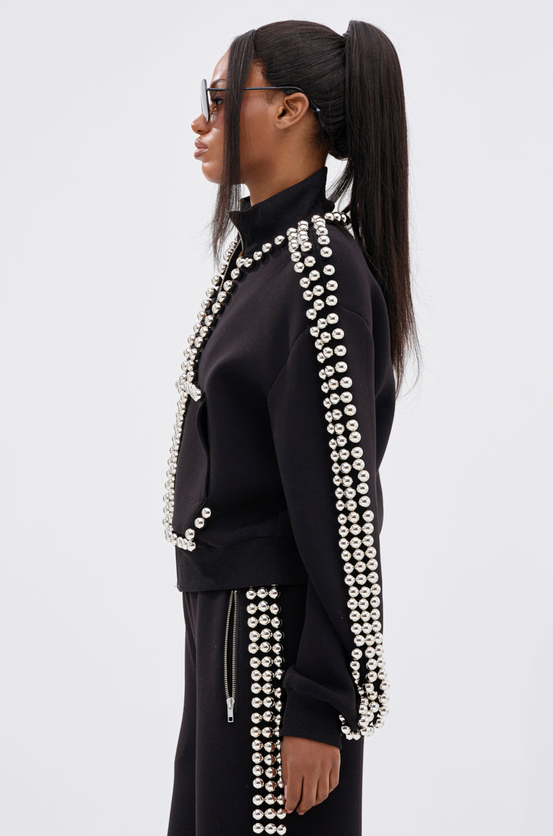 A woman with long straight hair in a high ponytail wears the BALLS TO THE WALL ZIP UP SWEATSHIRT IN BLACK with oversized silver ball details and black sunglasses, standing in profile against a plain white background.
