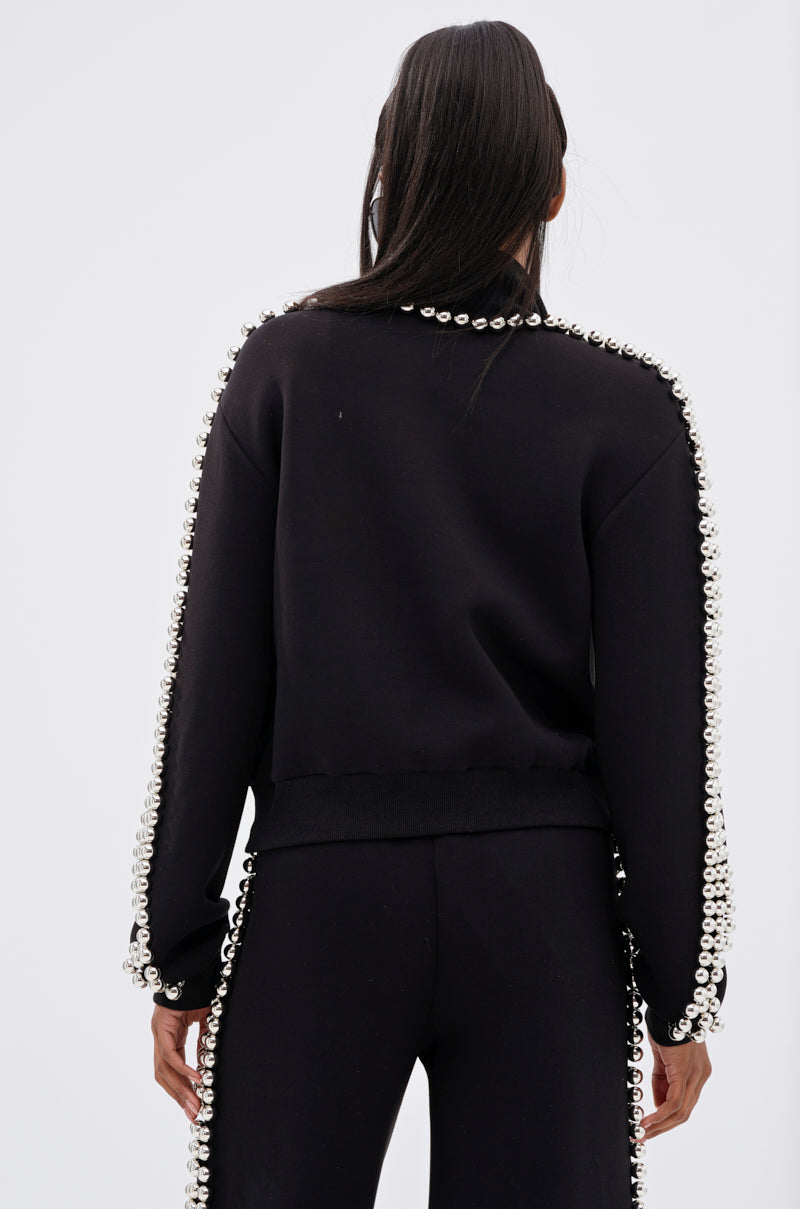 A person with long dark hair, seen from behind, wears the BALLS TO THE WALL ZIP UP SWEATSHIRT IN BLACK, featuring oversized silver ball embellishments on the sleeves, standing against a plain light background.
