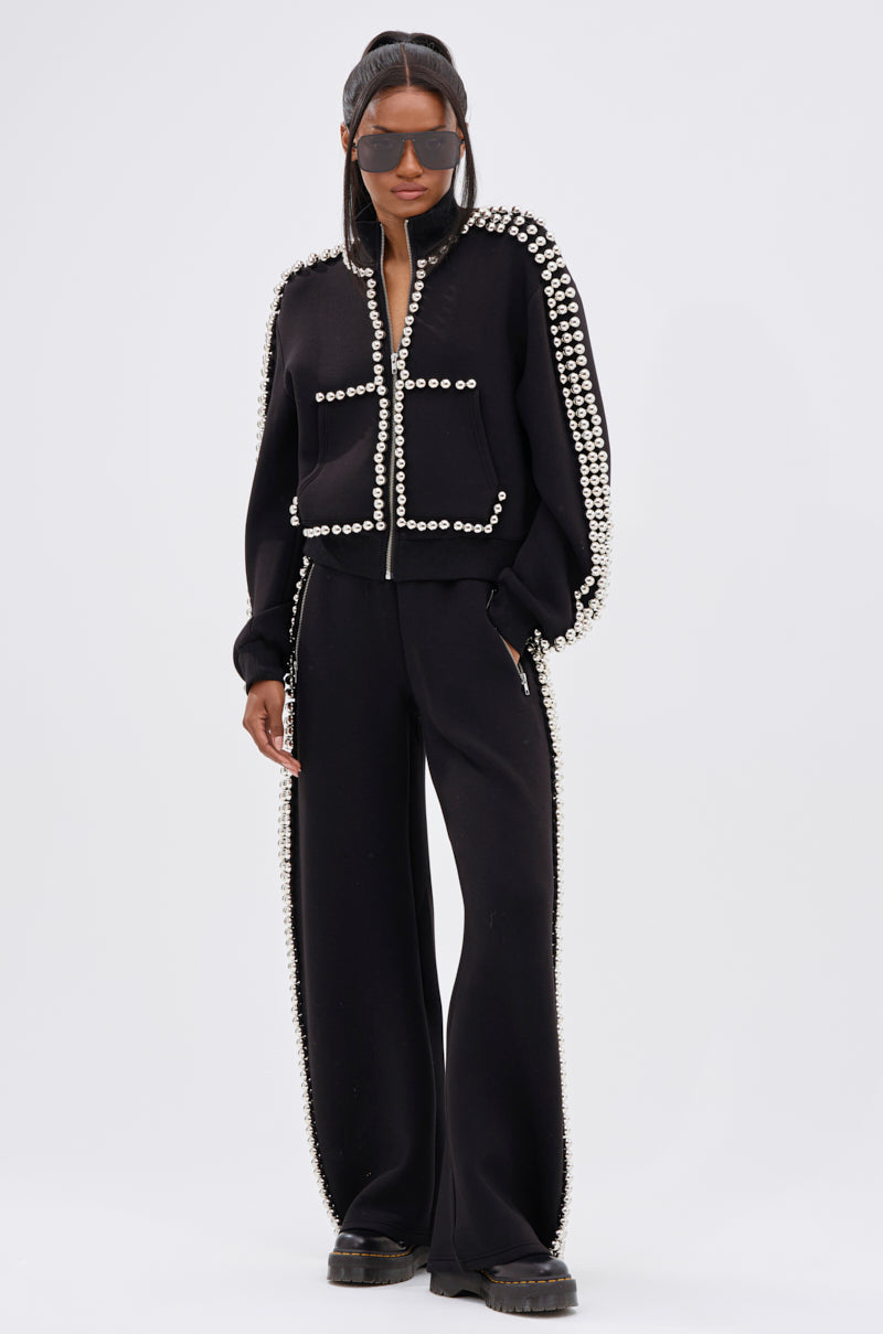 A person in oversized sunglasses and a black two-piece, with pearl-like details similar to the BALLS TO THE WALL ZIP UP SWEATSHIRT IN BLACK, stands confidently against a plain white background.