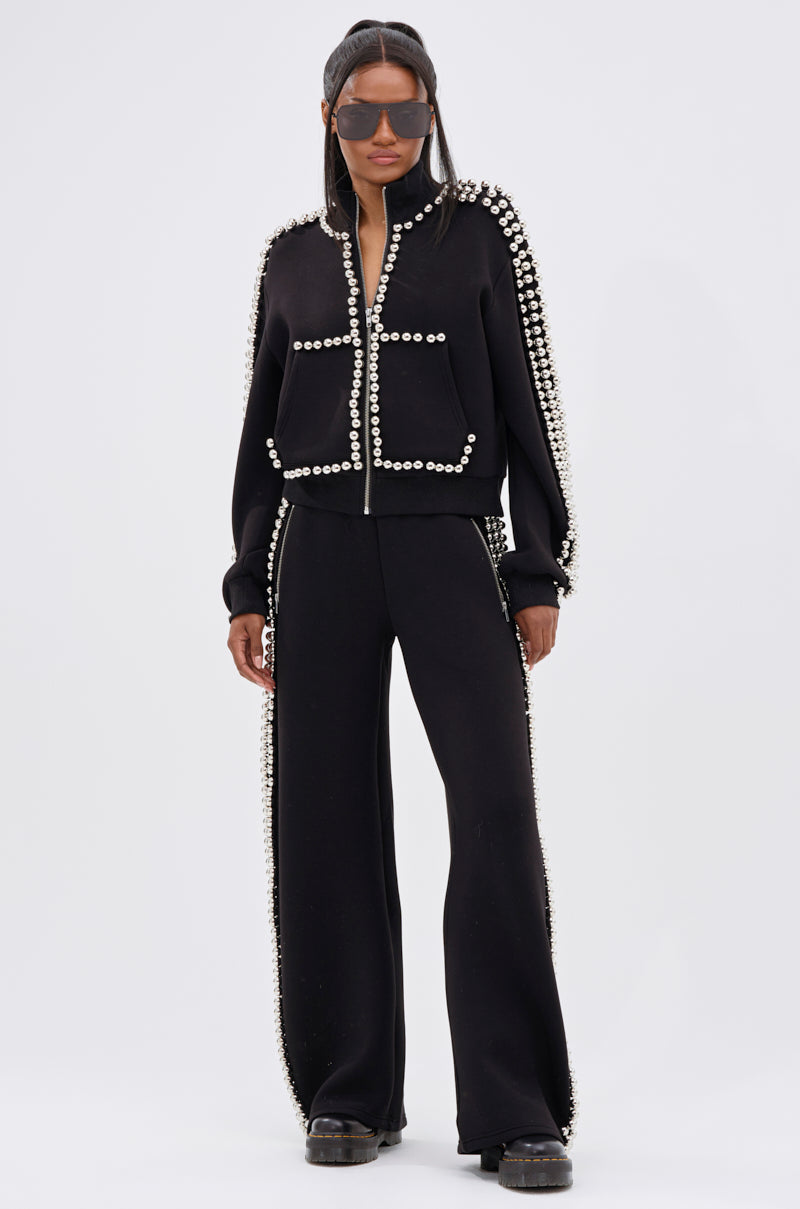 A woman stands confidently in the BALLS TO THE WALL ZIP UP SWEATSHIRT IN BLACK, paired with matching pearl-embellished pants, black platform shoes, and oversized sunglasses against a plain light background.