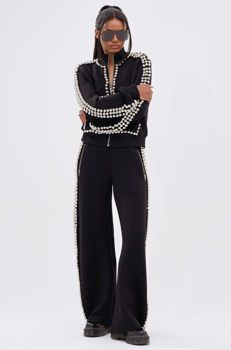 A woman stands with arms crossed, wearing chunky black shoes and the BALLS TO THE WALL ZIP UP SWEATSHIRT IN BLACK, which features rows of large silver studs and matching studded trim on both the jacket and wide-leg pants.