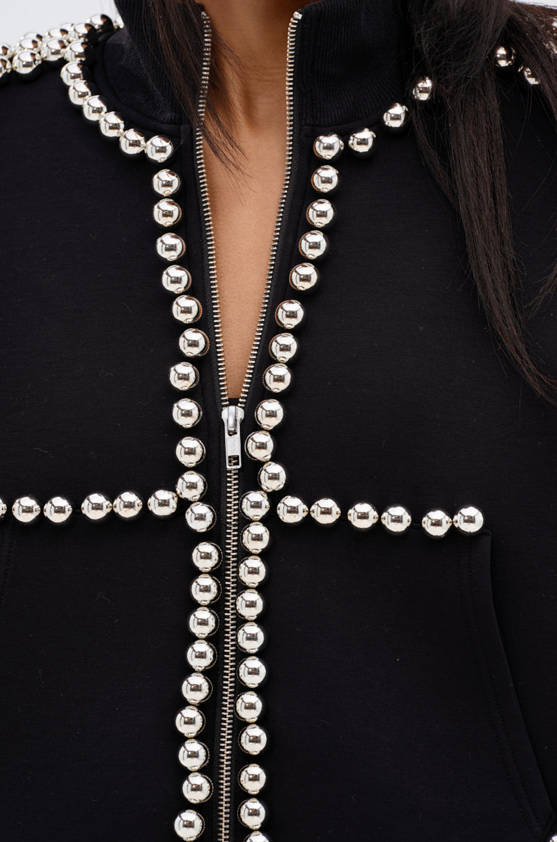 Close-up of a person wearing the BALLS TO THE WALL ZIP UP SWEATSHIRT IN BLACK, adorned with oversized silver ball embellishments in a striking cross pattern on the front; the person's face is not visible.