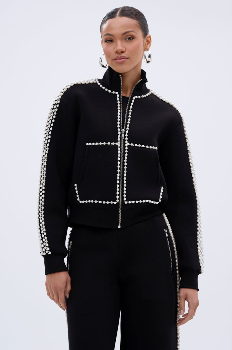A woman with her hair pulled back and wearing large silver hoop earrings stands against a plain background in the PEARL GIRL ZIP UP SWEATSHIRT, embellished with rows of white pearls along its edges and seams.