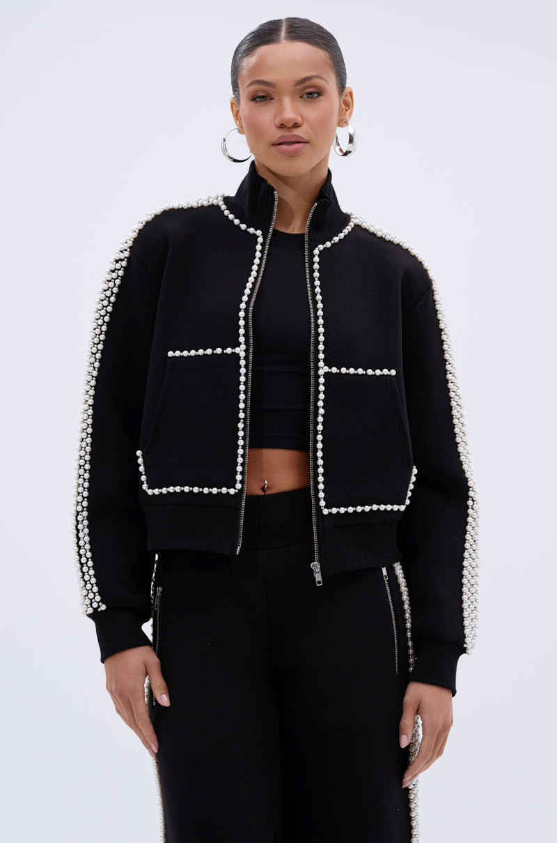A woman with slicked-back hair stands confidently against a plain, light background, wearing the PEARL GIRL ZIP UP SWEATSHIRT and matching pants adorned with rows of white pearl-like beads.