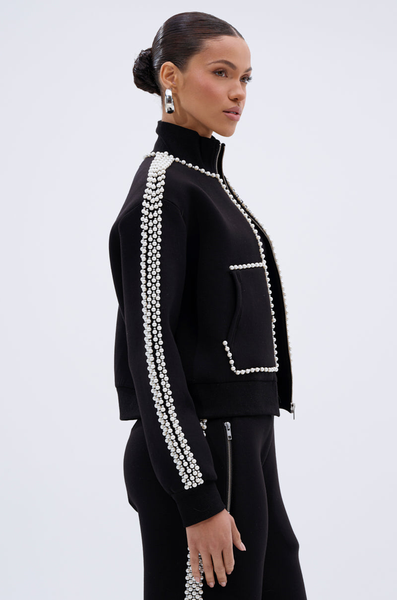 A woman with her hair in a sleek bun models the PEARL GIRL ZIP UP SWEATSHIRT, black with white pearl-like beads on the sleeves and pocket edges, paired with matching pants against a plain light background.