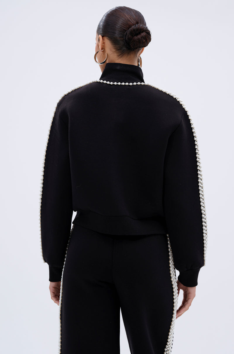 照片中,一位梳着利落发髻的女子背对着镜头,背景是纯浅色的珍珠女孩拉链卫衣和配套的黑色长裤,裤子的接缝处饰有珍珠。