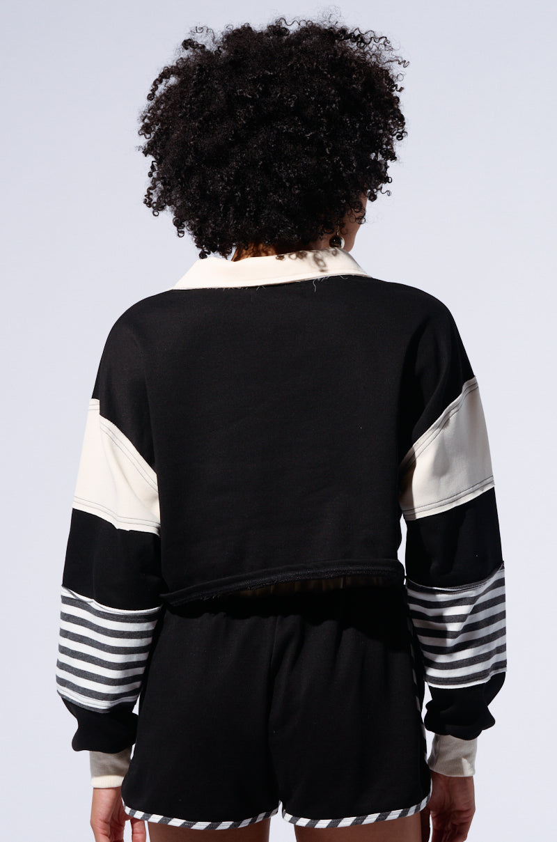A person with curly hair stands facing away, wearing an ON MY MIND CROP SWEATSHIRT and matching shorts. The black and white cotton-blend outfit boasts a color block pattern with horizontal stripes on the sleeves and shorts, creating a cohesive and stylish look. The background is plain and light-colored.