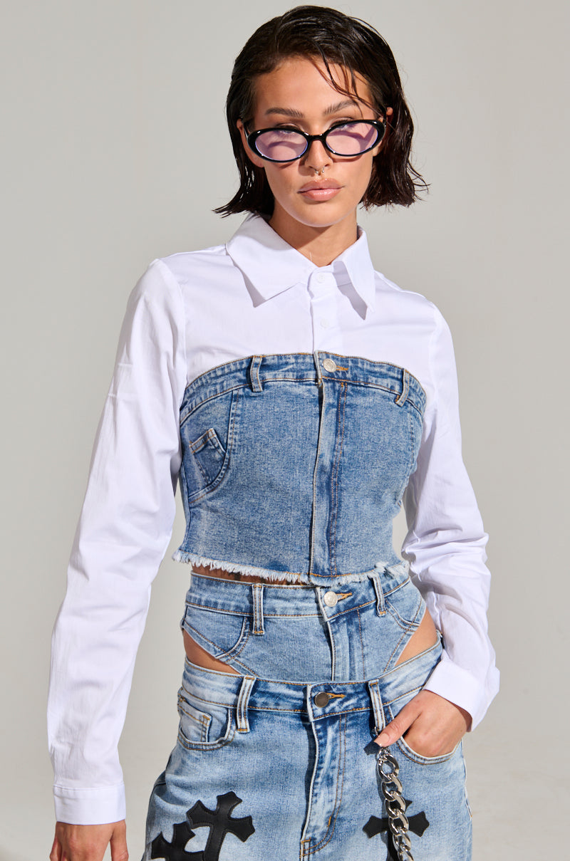 A person wearing a distinctive SITTING PRETTY POPLIN & DENIM LONG SLEEVE TOP, which features a white poplin button-up shirt with a cropped denim top overlay, paired with high-waisted denim pants. They have short hair and are wearing futuristic sunglasses while standing confidently with one hand in their pocket.