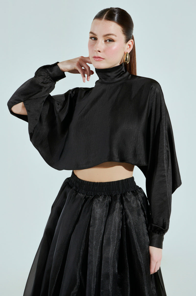 A woman poses against a plain background, wearing the TALIA SATIN MOCK NECK BLOUSE in black with dolman sleeves and a voluminous black skirt. Her hair is pulled back, and she wears large hoop earrings, exuding a thoughtful and confident expression.