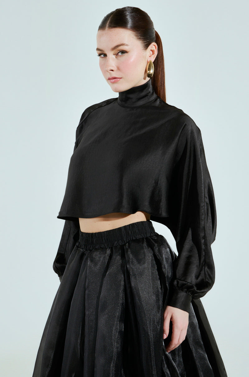 A person with long brown hair wears the TALIA SATIN MOCK NECK BLOUSE and a voluminous skirt, standing against a light background. They accessorize with large hoop earrings and gaze at the camera with a neutral expression.