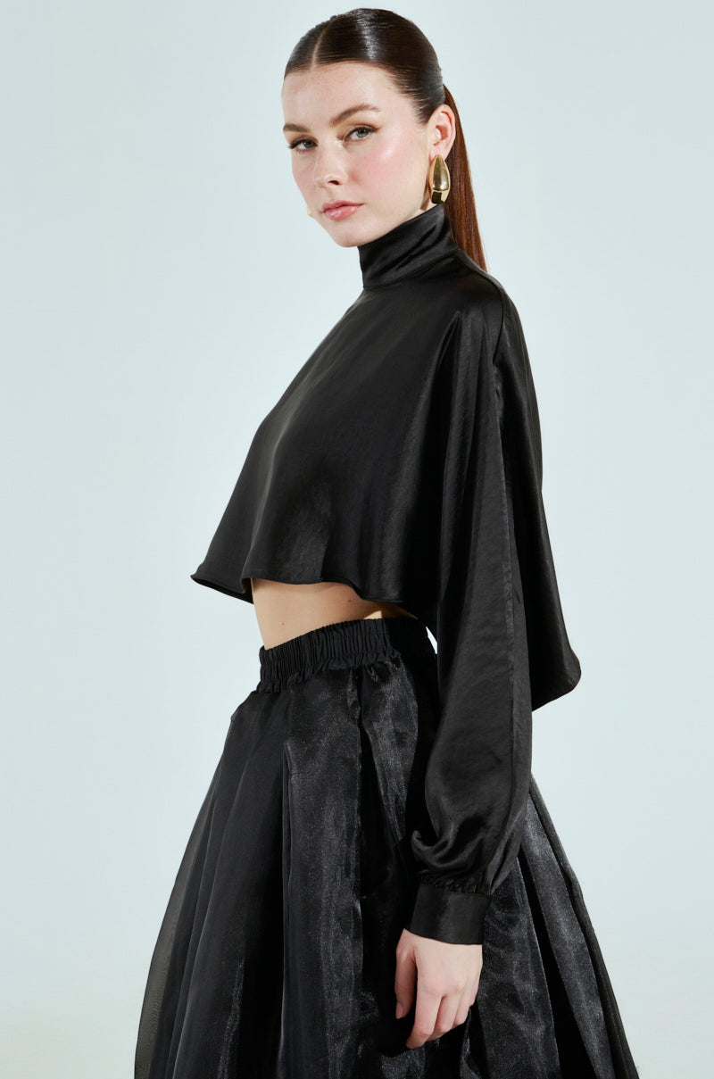 A woman poses sideways against a light background, wearing the TALIA SATIN MOCK NECK BLOUSE in black satin, featuring long dolman sleeves, paired with a flowing black skirt. Her hair is tied back.
