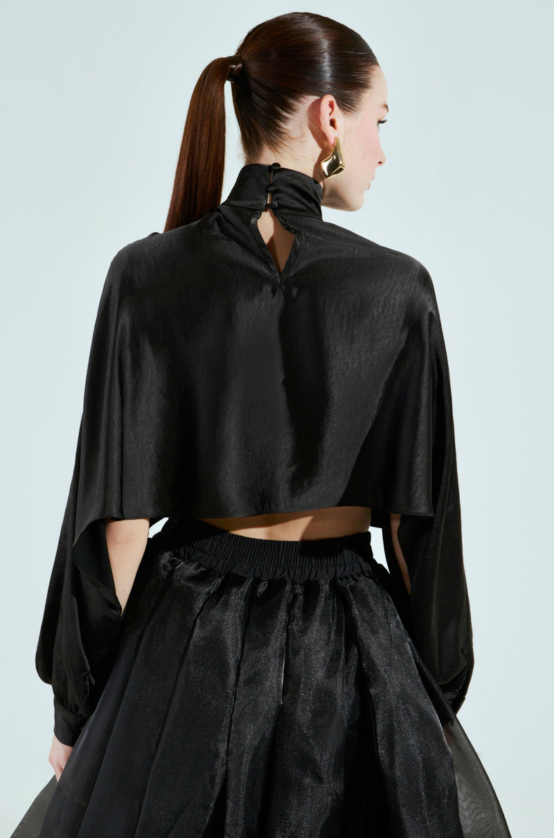 A person with a long ponytail is wearing a stylish black ensemble, featuring the TALIA SATIN MOCK NECK BLOUSE and a gown with a voluminous skirt. They are turned away, highlighting the intricate back details of the outfit.