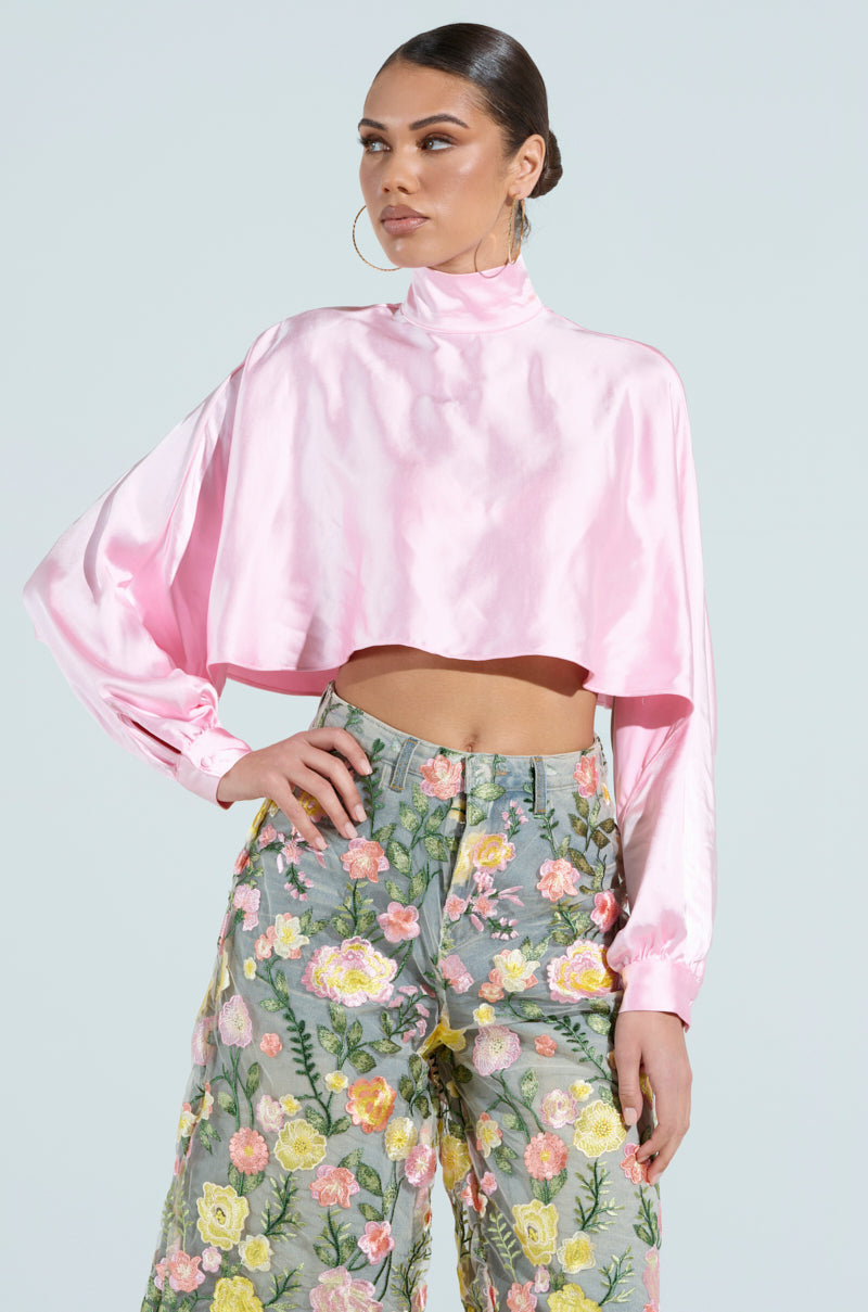 A woman models the TALIA SATIN MOCK NECK BLOUSE with a shiny pink cropped fit, paired with high-waisted pants featuring floral embroidery. Hoop earrings complement her sleek ponytail as she stands against a light gray background.