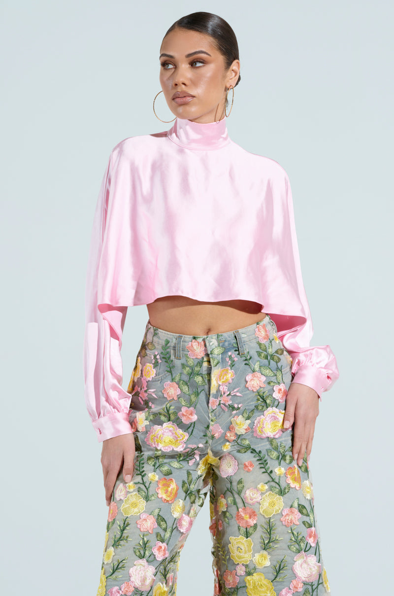 A woman poses against a light blue background wearing the TALIA SATIN MOCK NECK BLOUSE in pink with bishop sleeves, paired with gray floral high-waisted pants. She accessorizes with hoop earrings and has her hair pulled back.