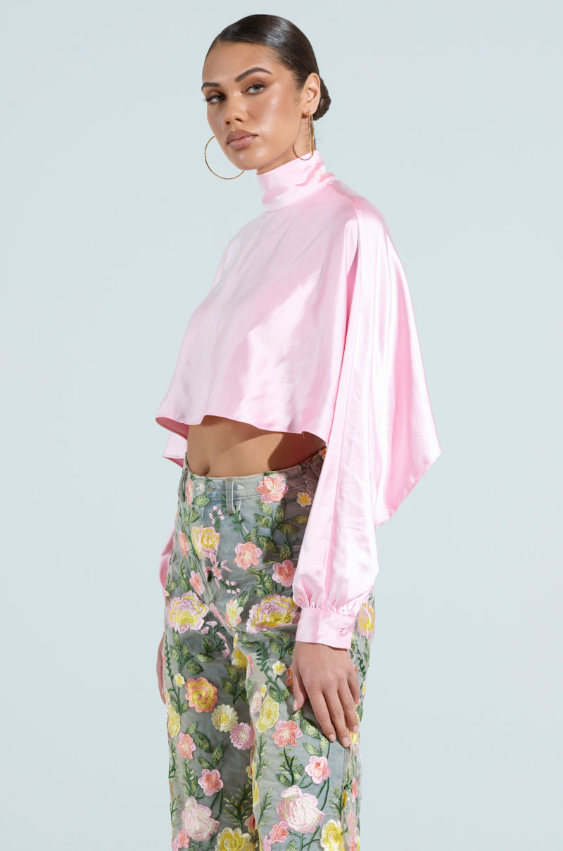 Someone is wearing the TALIA SATIN MOCK NECK BLOUSE in pink, a cropped satin top with bishop sleeves, paired with floral pants featuring large yellow, pink, and green flowers. The outfit is enhanced by large hoop earrings and styled brown hair against a plain light gray background.