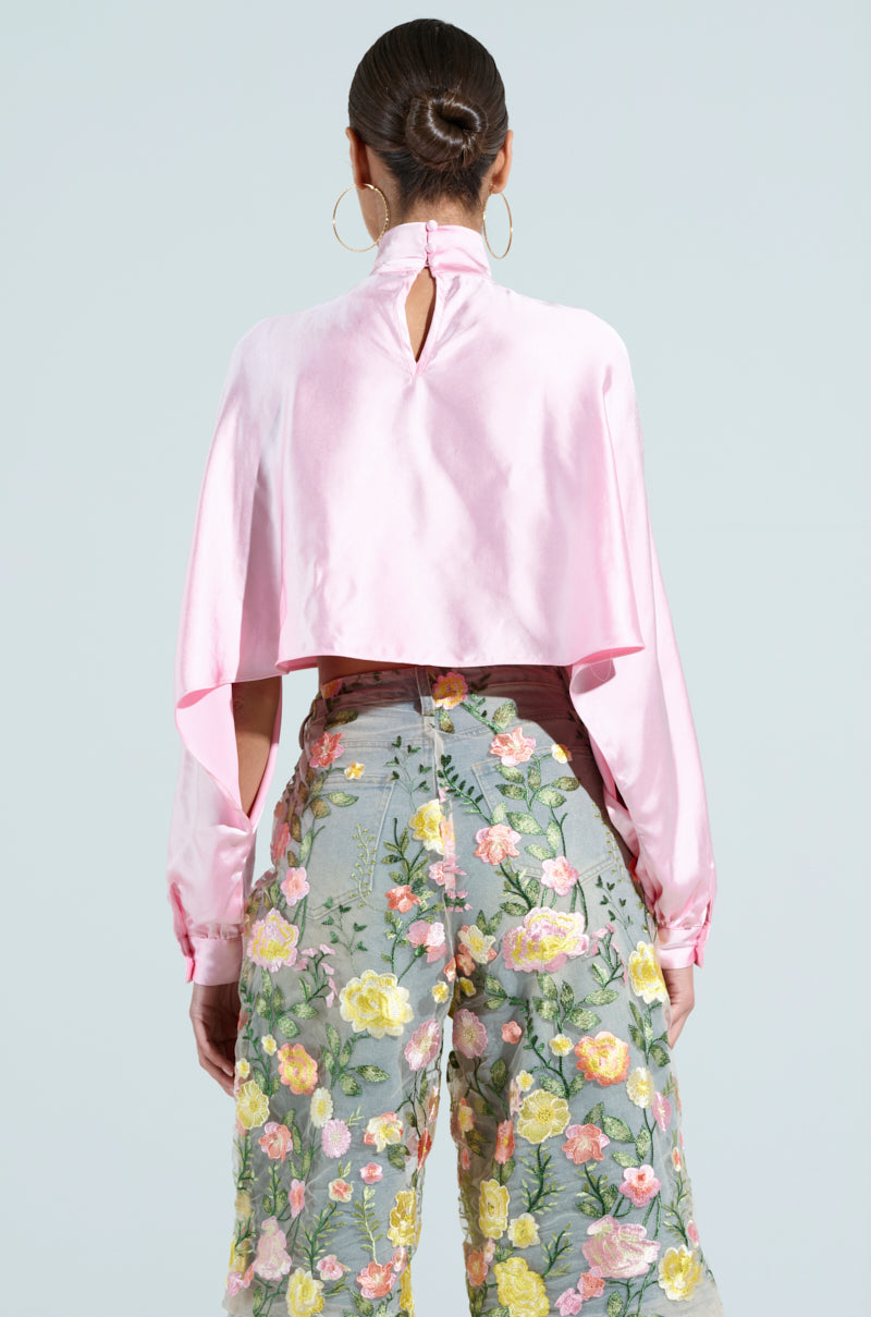 Against a light blue backdrop, someone with their hair in a bun wears the TALIA SATIN MOCK NECK BLOUSE in light pink, featuring bishop sleeves and a keyhole back. They're paired with sheer pants embellished with colorful embroidered flowers, exuding elegance.