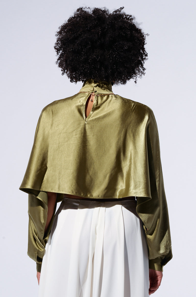 A person with curly hair is shown from the back wearing a Satin Mock Neck Blouse in Olive, featuring a keyhole opening and long sleeves, paired with white high-waisted pants. The background is plain and light-colored.