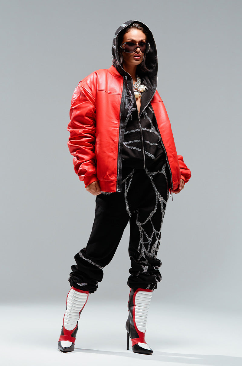 A person wearing a red leather jacket over a black hoodie, SPIDER SEQUIN DETAIL JOGGER pants, and red and white high-heeled boots. They also wear dark sunglasses and have a confident pose, looking stylish and modern against a plain backdrop.