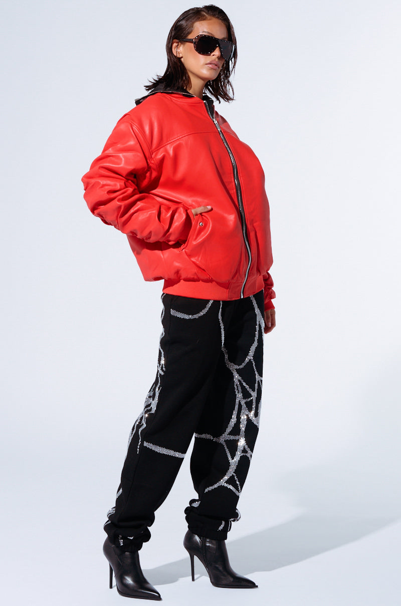 A person clad in a large red jacket, SPIDER SEQUIN DETAIL JOGGER pants adorned with white web-like designs, black high-heeled ankle boots, and dark sunglasses strikes a confident pose against a plain white background.