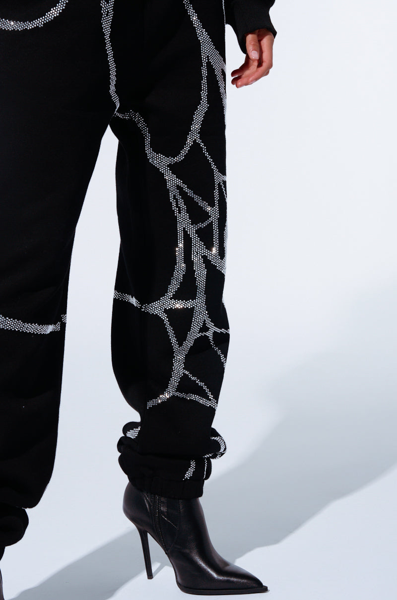 A person is wearing the SPIDER SEQUIN DETAIL JOGGER pants, featuring black fabric with white geometric patterns, paired with sleek black high-heeled boots adorned with subtle rhinestone detailing. Only the lower half of their body and part of their arm are visible against a plain, light background.