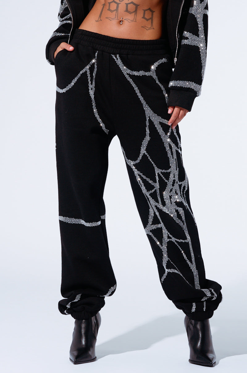 A person is wearing black joggers adorned with an abstract, web-like silver pattern, reminiscent of the SPIDER SEQUIN DETAIL JOGGER. The high-rise design pairs perfectly with black high-heeled boots, and they have their hands in the pockets. A tattoo reading "1999" is visible on their midriff.