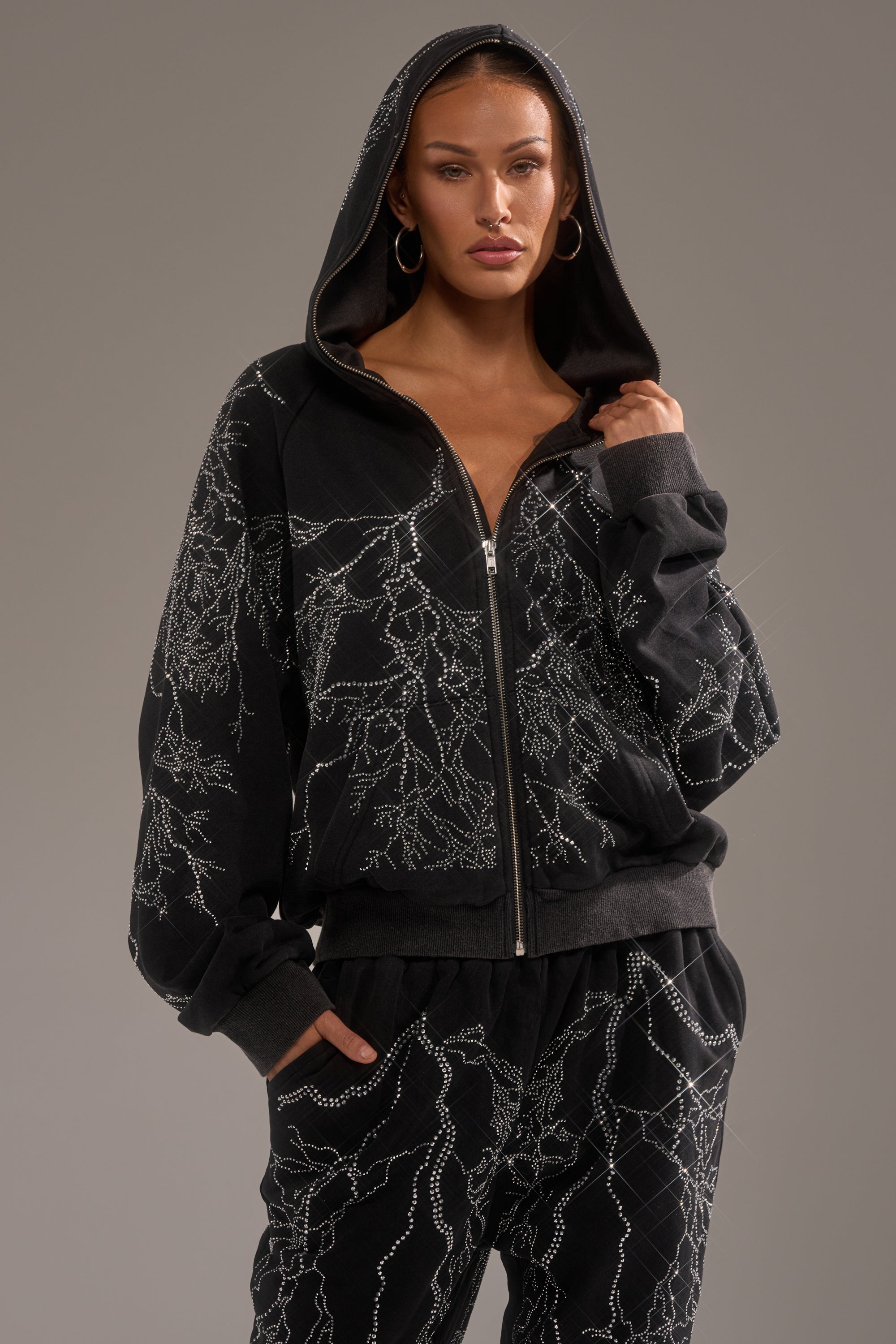 A woman confidently wears the ELECTRIC LOVE RHINESTONE ZIP UP SWEATSHIRT with white abstract line patterns, hood up, standing against a neutral background. 0