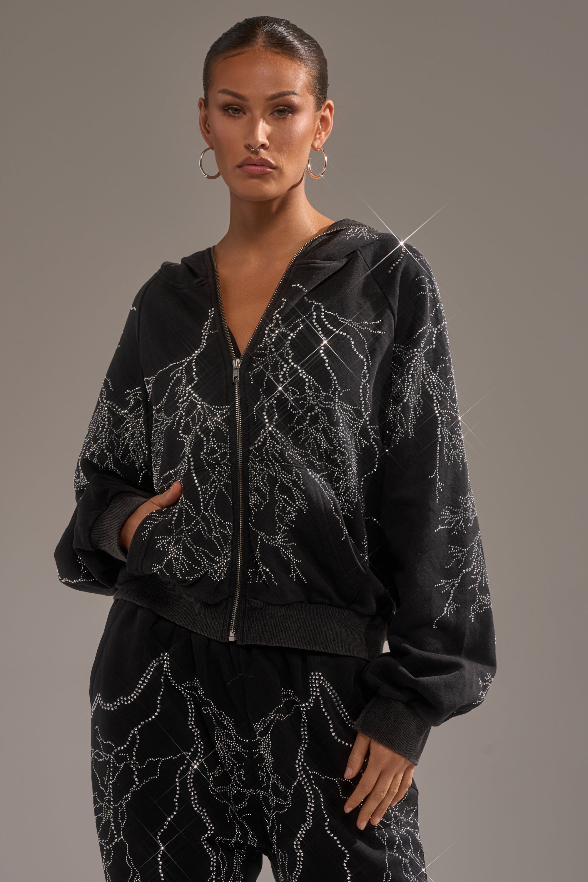 A woman wearing the ELECTRIC LOVE RHINESTONE ZIP UP SWEATSHIRT with dazzling, lightning-inspired rhinestones stands with one hand in her pocket against a neutral background. 1
