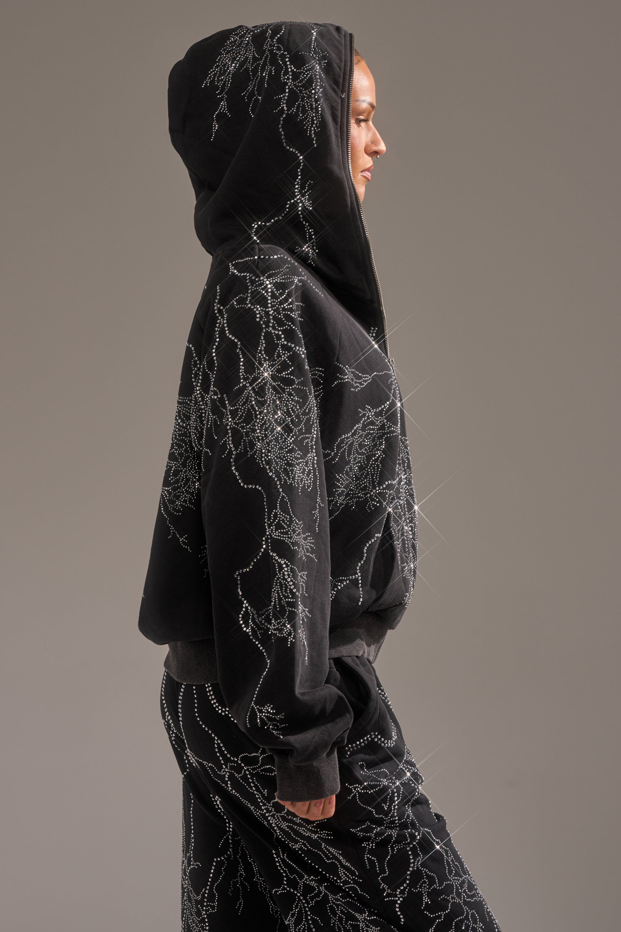 A person in profile models the ELECTRIC LOVE RHINESTONE ZIP UP SWEATSHIRT and matching pants, both featuring sparkling white branch-like patterns that stand out against a neutral gray background. 2